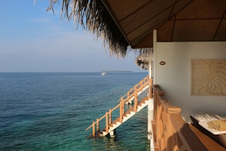 A serene view from a thatched-roof hut overlooking a vast expanse of blue ocean. A wooden staircase leads directly into the clear waters, and a small boat is visible in the distance. Inside the hut, there is a wall decor and a comfortable seating area bathed in sunlight.
