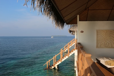 A serene view from a thatched-roof hut overlooking a vast expanse of blue ocean. A wooden staircase leads directly into the clear waters, and a small boat is visible in the distance. Inside the hut, there is a wall decor and a comfortable seating area bathed in sunlight.