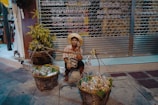 A street vendor sits on the sidewalk, dressed in a traditional hat and colorful shirt. They are surrounded by baskets filled with goods, possibly snacks or produce, ready for sale. The background consists of a closed metal storefront with signage written in a local language, and a potted plant adds greenery to the scene.