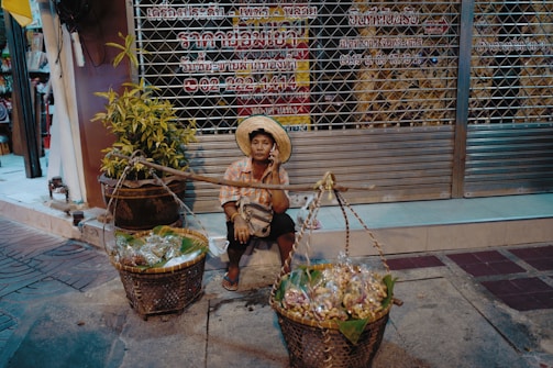 A street vendor sits on the sidewalk, dressed in a traditional hat and colorful shirt. They are surrounded by baskets filled with goods, possibly snacks or produce, ready for sale. The background consists of a closed metal storefront with signage written in a local language, and a potted plant adds greenery to the scene.