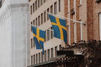Two Swedish flags are displayed on flagpoles attached to the side of a brick building. The facade of the building features multiple windows, and another structure wrapped in white material is visible in the background. The flags are blue with a yellow Nordic cross.