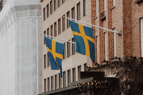 Two Swedish flags are displayed on flagpoles attached to the side of a brick building. The facade of the building features multiple windows, and another structure wrapped in white material is visible in the background. The flags are blue with a yellow Nordic cross.