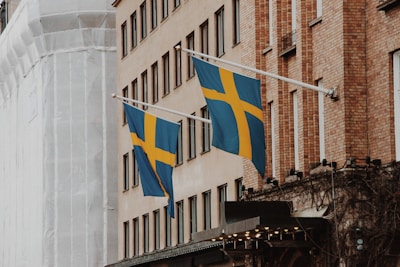 Two Swedish flags are displayed on flagpoles attached to the side of a brick building. The facade of the building features multiple windows, and another structure wrapped in white material is visible in the background. The flags are blue with a yellow Nordic cross.