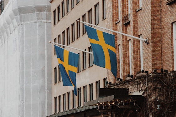 Two Swedish flags are displayed on flagpoles attached to the side of a brick building. The facade of the building features multiple windows, and another structure wrapped in white material is visible in the background. The flags are blue with a yellow Nordic cross.