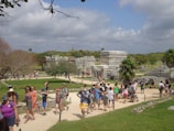 A bustling archaeological site with a group of people walking along a pathway surrounded by ancient stone structures. The scene includes vibrant greenery and a partly cloudy sky. The area has a lively atmosphere with tourists exploring the historic ruins.