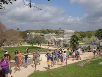 A bustling archaeological site with a group of people walking along a pathway surrounded by ancient stone structures. The scene includes vibrant greenery and a partly cloudy sky. The area has a lively atmosphere with tourists exploring the historic ruins.