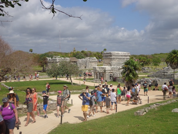 A bustling archaeological site with a group of people walking along a pathway surrounded by ancient stone structures. The scene includes vibrant greenery and a partly cloudy sky. The area has a lively atmosphere with tourists exploring the historic ruins.