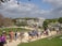A bustling archaeological site with a group of people walking along a pathway surrounded by ancient stone structures. The scene includes vibrant greenery and a partly cloudy sky. The area has a lively atmosphere with tourists exploring the historic ruins.