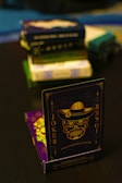 A playing card featuring a stylized sugar skull design, labeled 'Joker', is prominently displayed in the foreground. The card has an intricate design with a hat on the skull and decorative elements in gold. In the background, multiple stacks of board games or boxes are slightly out of focus, placed on a dark tabletop.