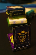 A playing card featuring a stylized sugar skull design, labeled 'Joker', is prominently displayed in the foreground. The card has an intricate design with a hat on the skull and decorative elements in gold. In the background, multiple stacks of board games or boxes are slightly out of focus, placed on a dark tabletop.