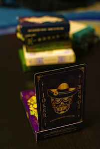 A playing card featuring a stylized sugar skull design, labeled 'Joker', is prominently displayed in the foreground. The card has an intricate design with a hat on the skull and decorative elements in gold. In the background, multiple stacks of board games or boxes are slightly out of focus, placed on a dark tabletop.