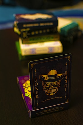 A playing card featuring a stylized sugar skull design, labeled 'Joker', is prominently displayed in the foreground. The card has an intricate design with a hat on the skull and decorative elements in gold. In the background, multiple stacks of board games or boxes are slightly out of focus, placed on a dark tabletop.