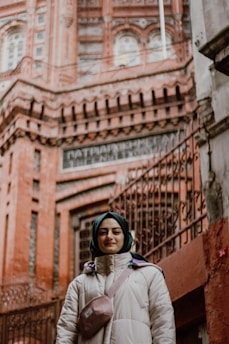 A person wearing a hijab and a winter coat stands in front of a historical brick building with ornate architectural details and iron railings.