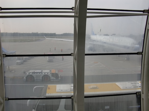 An airport scene viewed through large glass windows, showing a runway area with an airplane partially visible in the background. Ground support vehicles and airport staff in reflective vests are visible on the tarmac. The environment appears overcast with a subdued atmosphere.