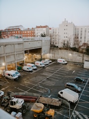 A large urban parking lot is partially filled with several white delivery vans, a few black cars, and construction machinery, including a digger and a dump truck with dirt. Surrounding the lot are industrial and residential buildings, some with graffiti and red rooftops. The sky is overcast, suggesting a gray day.