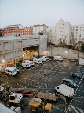 A large urban parking lot is partially filled with several white delivery vans, a few black cars, and construction machinery, including a digger and a dump truck with dirt. Surrounding the lot are industrial and residential buildings, some with graffiti and red rooftops. The sky is overcast, suggesting a gray day.