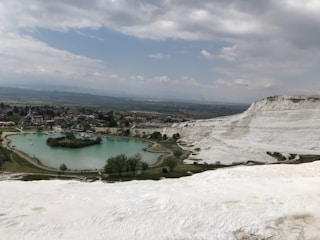 A scenic view of white travertine terraces cascading down a hillside with a clear turquoise pond in the foreground. The landscape is dotted with green vegetation and a small settlement is visible beyond the pond. The sky is overcast with thick clouds, adding a serene atmosphere to the scene.