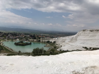 A scenic view of white travertine terraces cascading down a hillside with a clear turquoise pond in the foreground. The landscape is dotted with green vegetation and a small settlement is visible beyond the pond. The sky is overcast with thick clouds, adding a serene atmosphere to the scene.