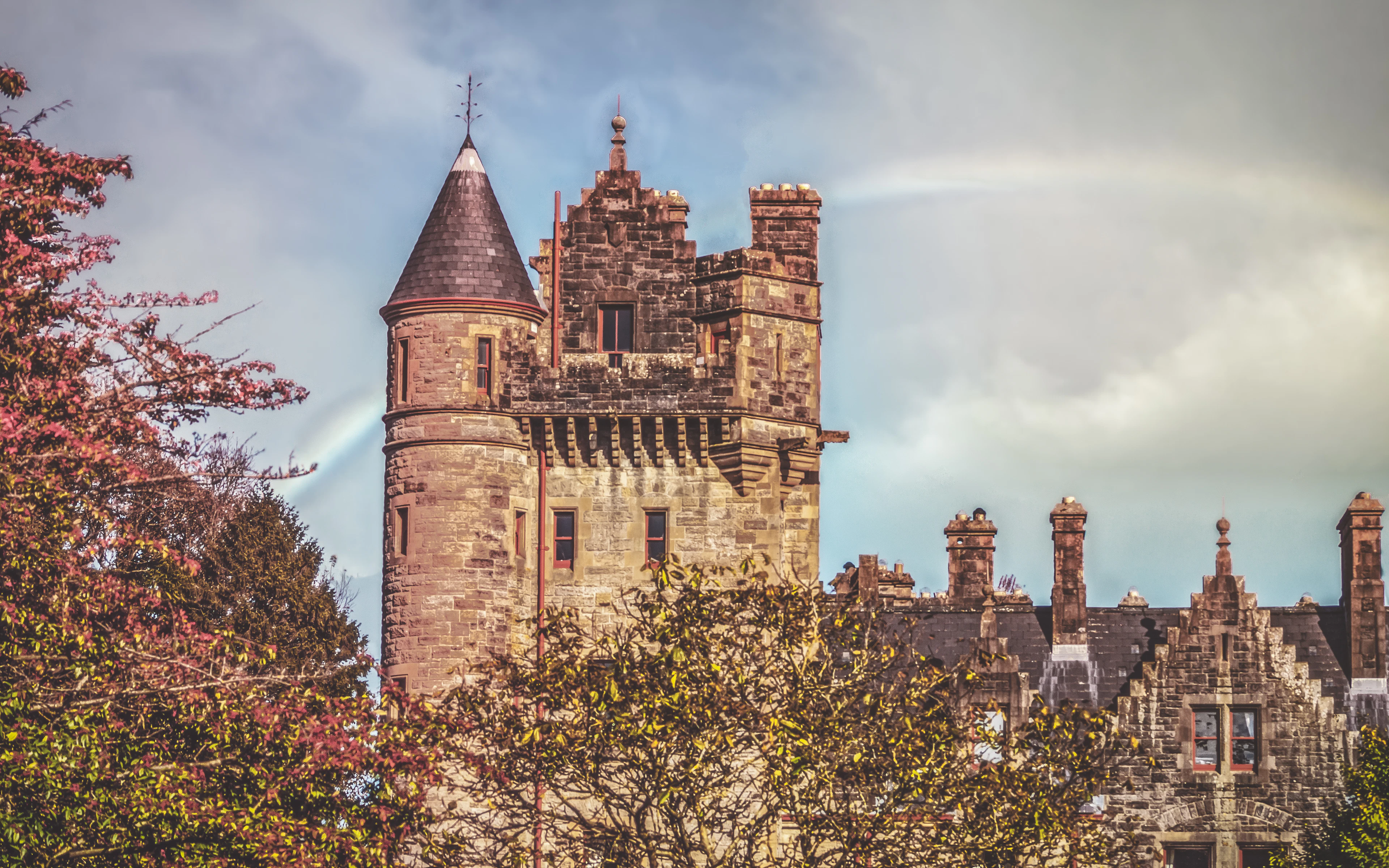 A turreted stone mansion in Scottish Baronial style framed by autumn trees, with a rainbow overhead.