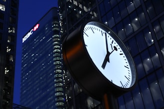 A large clock is prominently displayed in the foreground against the backdrop of a modern cityscape. The clock face is round, with sleek black hour and minute hands on a white background. In the background, skyscrapers with reflective glass facades are illuminated, with one building having visible branding.