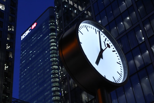 A large clock is prominently displayed in the foreground against the backdrop of a modern cityscape. The clock face is round, with sleek black hour and minute hands on a white background. In the background, skyscrapers with reflective glass facades are illuminated, with one building having visible branding.