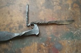 A handcrafted twisted metal knife with an ornate handle is placed on a rustic, textured wooden surface. The blade and handle show signs of artisanal craftsmanship, with intricate twists and a spiraling design.