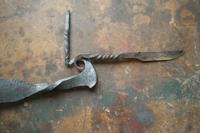 A handcrafted twisted metal knife with an ornate handle is placed on a rustic, textured wooden surface. The blade and handle show signs of artisanal craftsmanship, with intricate twists and a spiraling design.