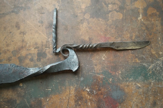 A handcrafted twisted metal knife with an ornate handle is placed on a rustic, textured wooden surface. The blade and handle show signs of artisanal craftsmanship, with intricate twists and a spiraling design.
