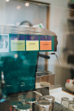 A coffee machine with colorful labels is set on a countertop, surrounded by several glass jars and metal containers. The machine has a shiny metallic finish with vibrant color-coded labels, indicating different coffee types.