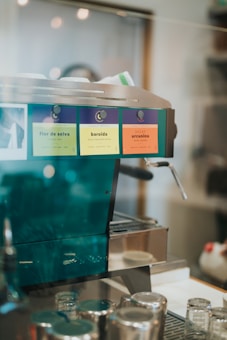 A coffee machine with colorful labels is set on a countertop, surrounded by several glass jars and metal containers. The machine has a shiny metallic finish with vibrant color-coded labels, indicating different coffee types.