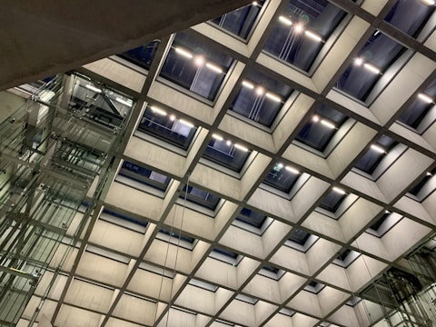 An architectural structure featuring a grid-like ceiling design with rectangular windows allowing light to enter. The ceiling is composed of concrete frames, and there is a visible elevator shaft with glass panels on the left side.