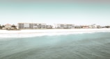 Several mid-rise buildings are lined up along a sandy beach, with the ocean in the foreground. The architecture is modern, and the scene has a serene and minimalistic atmosphere.