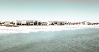 Several mid-rise buildings are lined up along a sandy beach, with the ocean in the foreground. The architecture is modern, and the scene has a serene and minimalistic atmosphere.