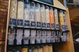 A series of transparent dispensers filled with various types of grains and seeds, each labeled with its name and price per kilogram. The containers include nut-free muesli, muesli base, porridge oats, jumbo oats, wheat flakes, rye flakes, barley flakes, red lentils, sunflower seeds, and whole-wheat penne. The setup is likely located in a shop or market with visible brick walls in the background.