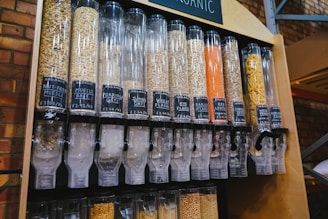 A series of transparent dispensers filled with various types of grains and seeds, each labeled with its name and price per kilogram. The containers include nut-free muesli, muesli base, porridge oats, jumbo oats, wheat flakes, rye flakes, barley flakes, red lentils, sunflower seeds, and whole-wheat penne. The setup is likely located in a shop or market with visible brick walls in the background.