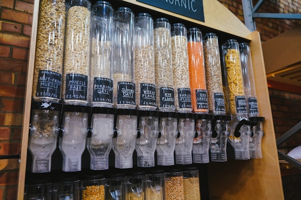 A series of transparent dispensers filled with various types of grains and seeds, each labeled with its name and price per kilogram. The containers include nut-free muesli, muesli base, porridge oats, jumbo oats, wheat flakes, rye flakes, barley flakes, red lentils, sunflower seeds, and whole-wheat penne. The setup is likely located in a shop or market with visible brick walls in the background.