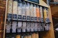 A series of transparent dispensers filled with various types of grains and seeds, each labeled with its name and price per kilogram. The containers include nut-free muesli, muesli base, porridge oats, jumbo oats, wheat flakes, rye flakes, barley flakes, red lentils, sunflower seeds, and whole-wheat penne. The setup is likely located in a shop or market with visible brick walls in the background.