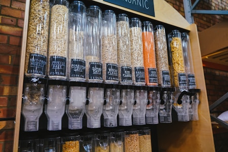A series of transparent dispensers filled with various types of grains and seeds, each labeled with its name and price per kilogram. The containers include nut-free muesli, muesli base, porridge oats, jumbo oats, wheat flakes, rye flakes, barley flakes, red lentils, sunflower seeds, and whole-wheat penne. The setup is likely located in a shop or market with visible brick walls in the background.