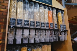 A series of transparent dispensers filled with various types of grains and seeds, each labeled with its name and price per kilogram. The containers include nut-free muesli, muesli base, porridge oats, jumbo oats, wheat flakes, rye flakes, barley flakes, red lentils, sunflower seeds, and whole-wheat penne. The setup is likely located in a shop or market with visible brick walls in the background.