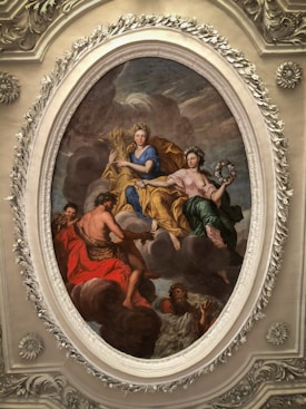 An ornate oval painting depicts several mythological figures amidst clouds. The central figure is a woman in blue holding a sheaf of wheat, symbolizing abundance. Another figure in a green dress holds a wreath, while a muscular figure in red is depicted reaching out. The intricate frame surrounding the painting features floral motifs.