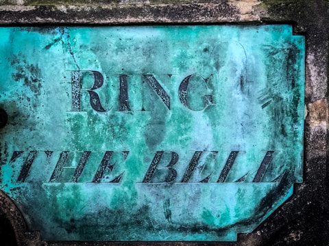 A weathered, teal-colored metal plaque with the words 'RING THE BELL' embossed in large, bold letters. The plaque has visible signs of aging, including rust and scratches, and is mounted on a stone surface.