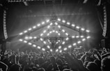 A concert scene with a large crowd of people raising their hands, enjoying live music. A stage features a geometric light structure with bright, square lights in a circular pattern. The environment suggests an energetic and lively atmosphere, with smoke or mist enhancing the light effects.