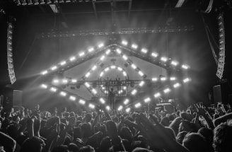 A concert scene with a large crowd of people raising their hands, enjoying live music. A stage features a geometric light structure with bright, square lights in a circular pattern. The environment suggests an energetic and lively atmosphere, with smoke or mist enhancing the light effects.