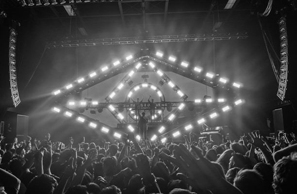 A concert scene with a large crowd of people raising their hands, enjoying live music. A stage features a geometric light structure with bright, square lights in a circular pattern. The environment suggests an energetic and lively atmosphere, with smoke or mist enhancing the light effects.