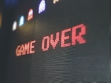 The image displays a pixelated 'Game Over' screen with the text prominently centered in red. Above the text, there are small, colorful pixelated characters resembling those from classic arcade games against a dark background.