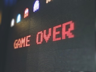 The image displays a pixelated 'Game Over' screen with the text prominently centered in red. Above the text, there are small, colorful pixelated characters resembling those from classic arcade games against a dark background.