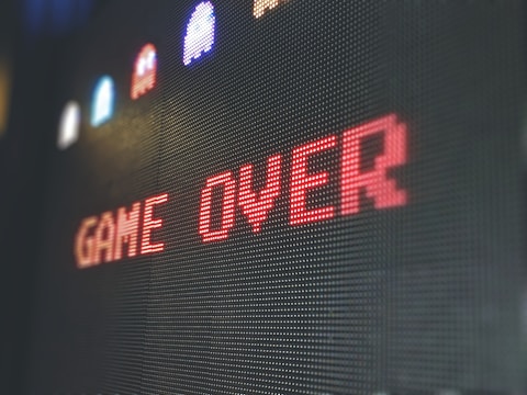 The image displays a pixelated 'Game Over' screen with the text prominently centered in red. Above the text, there are small, colorful pixelated characters resembling those from classic arcade games against a dark background.