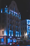 A multi-story building with architectural features illuminated by blue lighting, creating a striking contrast against the night sky. The facade displays the name 'Hotel Timisoara' in illuminated letters. The ground floor includes commercial establishments, with a recognizable coffee shop logo visible. Vehicles are parked or moving along the street in front.