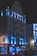 A multi-story building with architectural features illuminated by blue lighting, creating a striking contrast against the night sky. The facade displays the name 'Hotel Timisoara' in illuminated letters. The ground floor includes commercial establishments, with a recognizable coffee shop logo visible. Vehicles are parked or moving along the street in front.