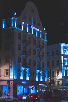 A multi-story building with architectural features illuminated by blue lighting, creating a striking contrast against the night sky. The facade displays the name 'Hotel Timisoara' in illuminated letters. The ground floor includes commercial establishments, with a recognizable coffee shop logo visible. Vehicles are parked or moving along the street in front.